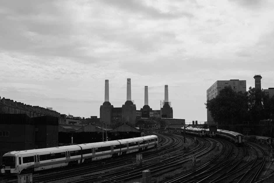 Black and white image of a railway yard with multiple train tracks curving toward a skyline featuring the Battersea Power Station with four tall chimneys, located in the background. The scene includes