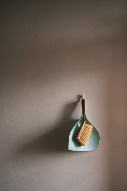 A light blue dustpan and a wooden-handled scrub brush with beige bristles hanging on a hook against a neutral grey wall, illustrating tools used in surface cleaning and dust removal as part of domesti